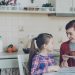 Young happy mother bringing tasty muffins to her cute daughter sitting at the table in kitchen waiting for breakfast at home
